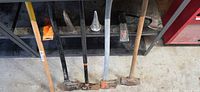 Four long-handled tools and spare handle leaning against shelf showing heads, plus wedges and hatchet visible on shelf