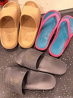 Three pairs of sandals laid out on floor: beige slides, gray slides with owl emblem, blue and pink flip-flops