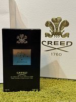 Front view of sealed Creed Aventus retail box beside Creed crest backdrop