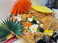 Pile of luau decorations showing orange honeycomb fan, green palm leaf fan, yellow spiral lantern, raffia matting and paper toucan cut-out