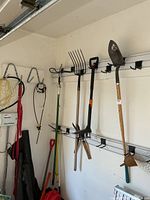 Garage wall rail holding pitchfork, shovel, pole pruner, clippers, hoe, fuel can and paddles in view