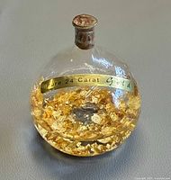 Front view of clear glass ball filled with gold flakes, cork top visible, label reads "Pure 24 Carat Gold"