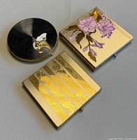 Front view of three compacts: round black enamel, square floral enamel, square striped floral engraving