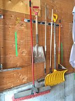 Overall wall view showing broom, shovel, two wooden-handled tools, ice scraper, and yellow snow shovel