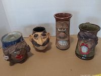 Front view of four face mugs showing sculpted features and glaze colors