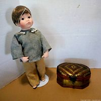 Full view of porcelain Oliver Twist doll on stand beside woven brass-trim box