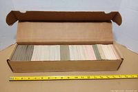 Open cardboard card storage box showing full rows of trading cards, ruler for scale