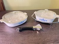 Two CorningWare casseroles with lids, chrome cradle, pan grabber