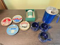 Overview of all nine items on table: four decorative cheese label plates, three blue speckled mugs, Starbucks travel cup in box, metallic blue pitcher with basket