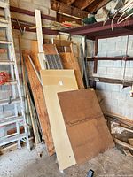 Stack of assorted plywood, particle board, OSB, drywall pieces, pegboard sheet, metal pipes and lumber leaning against garage wall