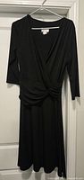Full view of black wrap dress hanging, showing front knot, V-neck, 3⁄4 sleeves, knee length