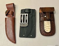 Front view of three sheaths: brown leather sheath, black Kydex sheath with clip, decorative brown leather pouch
