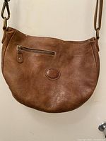 Front of Roots brown pebbled leather half-moon bag showing exterior zipper pocket and embossed logo patch