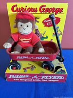 Front view of Curious George plush sitting in red Radio Flyer wagon on original packaging