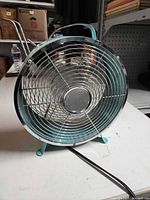 Front view of teal metal tabletop fan showing chrome grille and turquoise feet