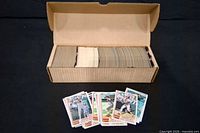 Open cardboard box holding large stack of baseball cards with several cards displayed in front