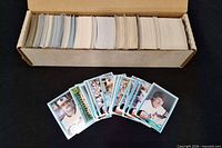 Open cardboard storage box showing large stack of baseball cards plus several cards fanned out in front