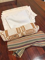 Stack of off-white tablecloths, white napkin, brown and white woven runner, multicolour striped runner