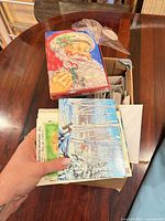 open cardboard box filled with assorted greeting cards and envelopes; Santa Christmas card box on top; hand holding winter scene card