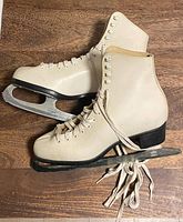 Side view of both skates showing white leather boots, laces, blades with toe picks, black heels