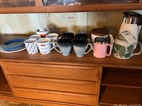 Group shot showing coffee pot, teapot, mugs, cups, canister, travel mug, juicer and plates on shelf