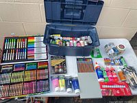 Overall view of art supplies laid out on table including plastic tote, art set trays, paint, glue, glitter and craft sticks