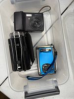 Front view of three cameras inside plastic bin: Samsung S730, black DVC camcorder, blue compact camera