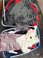 Two plastic bins filled with assorted clothing items including green patterned sweater, off-white scarf, red and grey garments, black pants, and plastic-wrapped textile