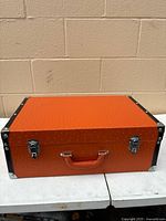 Closed orange suitcase front view showing latches, handle, black trim and corner protectors