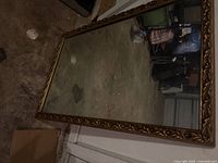 Full view of rectangular wall mirror leaning against wall, showing gold-tone ornate frame and mirror glass