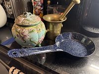 All three items grouped on countertop: brass mortar with pestle, ceramic cookie jar, speckled enamel pan