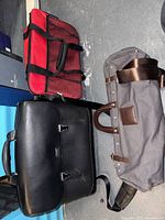 Overview of three travel bags: black Tumi briefcase, red carry bag, grey garment bag