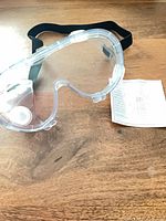 Opened sample safety goggles showing clear lens, black strap, side vent; instruction sheet visible