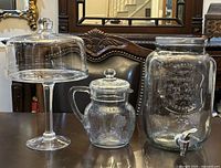 All three clear glass pieces—pitcher, cake stand with dome, beverage dispenser—group view