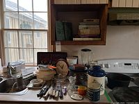 Overview of sink area showing blender, coffee maker, knives, utensils, clock, cookie jar, salt tin, plates, bowls, cast-iron skillet, tablecloth and placemats