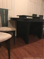 Side view of dark green dining table with one chair visible, showing table leg design and chair seat upholstery.