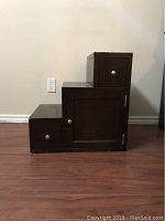 Photo of dark brown wooden side table or night stand with step design, showing three storage compartments with silver knobs, against a plain wall and hardwood floor.