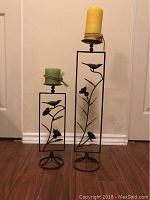 Photo of two metal candle holders with decorative bird and flower design; one taller with yellow candle, one shorter with green candle.