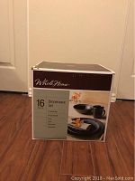 Box of the Whole Home 16 piece Dinnerware Set in black, showing brand, number of pieces, and image of the dishes included, box placed on a wooden floor.