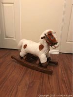 Plush rocking horse with white and brown spots and a wooden rocker base on a hardwood floor against a white door background.