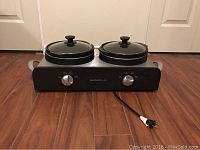 Electric Home Styles chafing dish with two covered pots on a stainless steel base with control knobs and a power cord, placed on a wooden floor in front of closed white doors.
