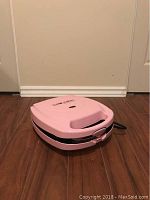 Side view photo of a pink Baby Cakes muffin maker sitting on a wooden floor against a beige wall.