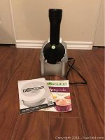 Silver and black Yonanas Maker kitchen appliance with power cord laid on a wooden floor along with user manual and recipe guide.