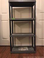 Front-facing photo of a black plastic garage shelf unit with four shelves shown standing against white double doors and brown wood floor. Unit appears solid and in excellent used condition.