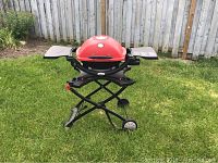 Weber grill with red lid on black foldable stand, side tables extended, placed outdoors on grass against wooden fence background