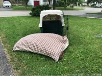Outdoor photo showing a beige and green plastic dog crate with an open metal door and a large checkered pillow placed on the grass in front of a house.