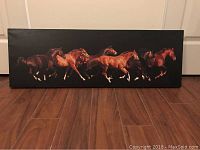 Canvas print displayed on wood floor, showing six running horses against a black background in landscape orientation.