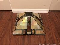Stained glass lamp shade lying on wooden floor showing top and side sides. Green, yellow, white, iridescent, and brown glass with metal framing.