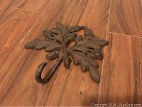 Close-up photo of a metal decorative wall hook shaped like an oak leaf with acorns on a wooden floor.