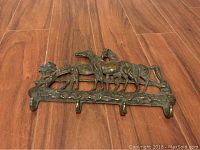 Photo of metal wall hook featuring four horses, showing front view on floor with wood grain background.